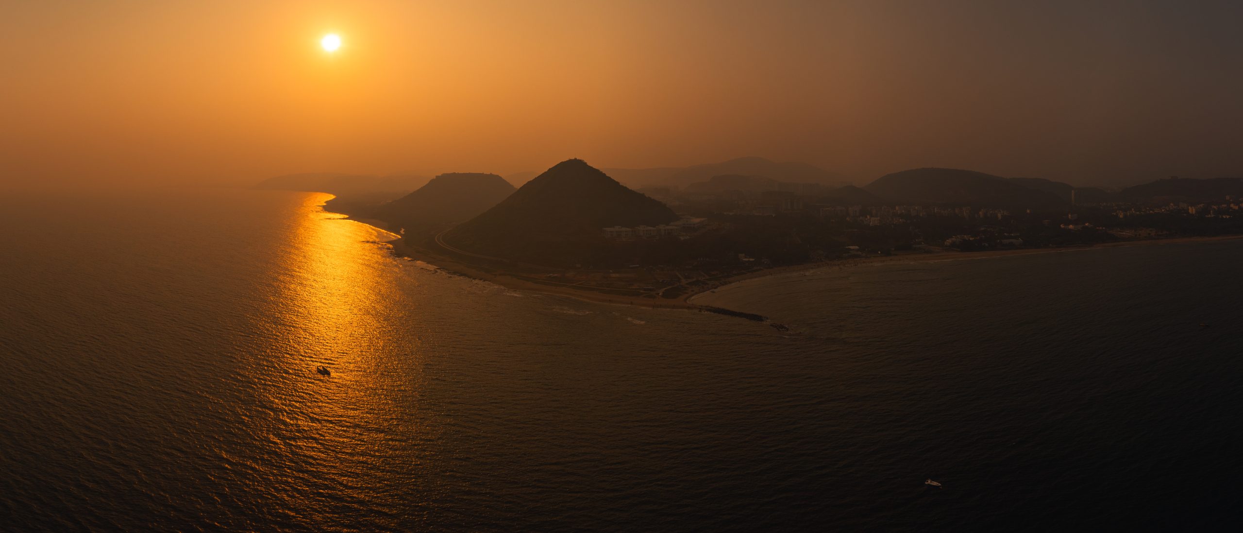 Aerial view of Vizag coastline at sunset, with golden sunlight reflecting over the Bay of Bengal and silhouetted hills along the shore.