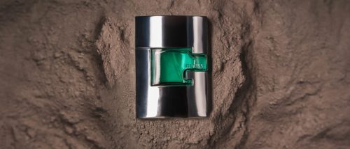 Green Guess perfume bottle embedded in textured brown sand with a focused light spill.