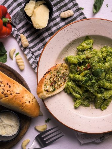 Plate of green pesto pasta with garlic bread, parmesan, and raw gnocchi on a rustic setup.