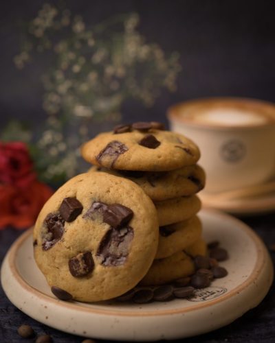 Stack of warm chocolate chunk cookies with coffee cup bokeh at The Funnel Hill Creamery.